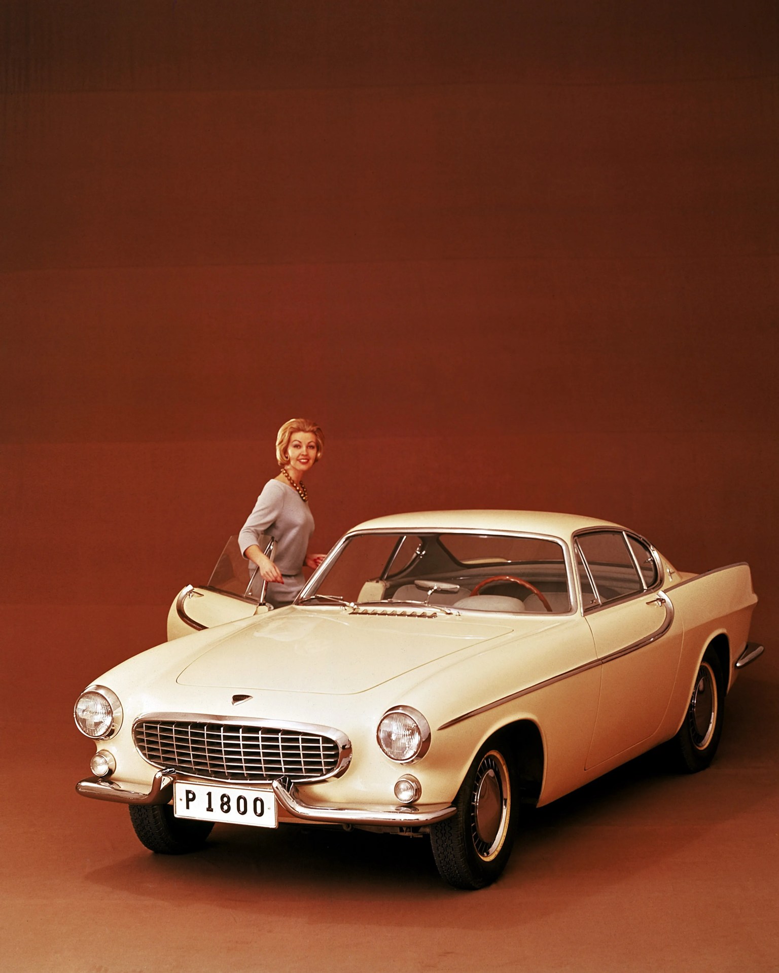 Cream-colored vintage Volvo P1800 coupe with chrome grille and bumpers, woman standing by open driver's door.