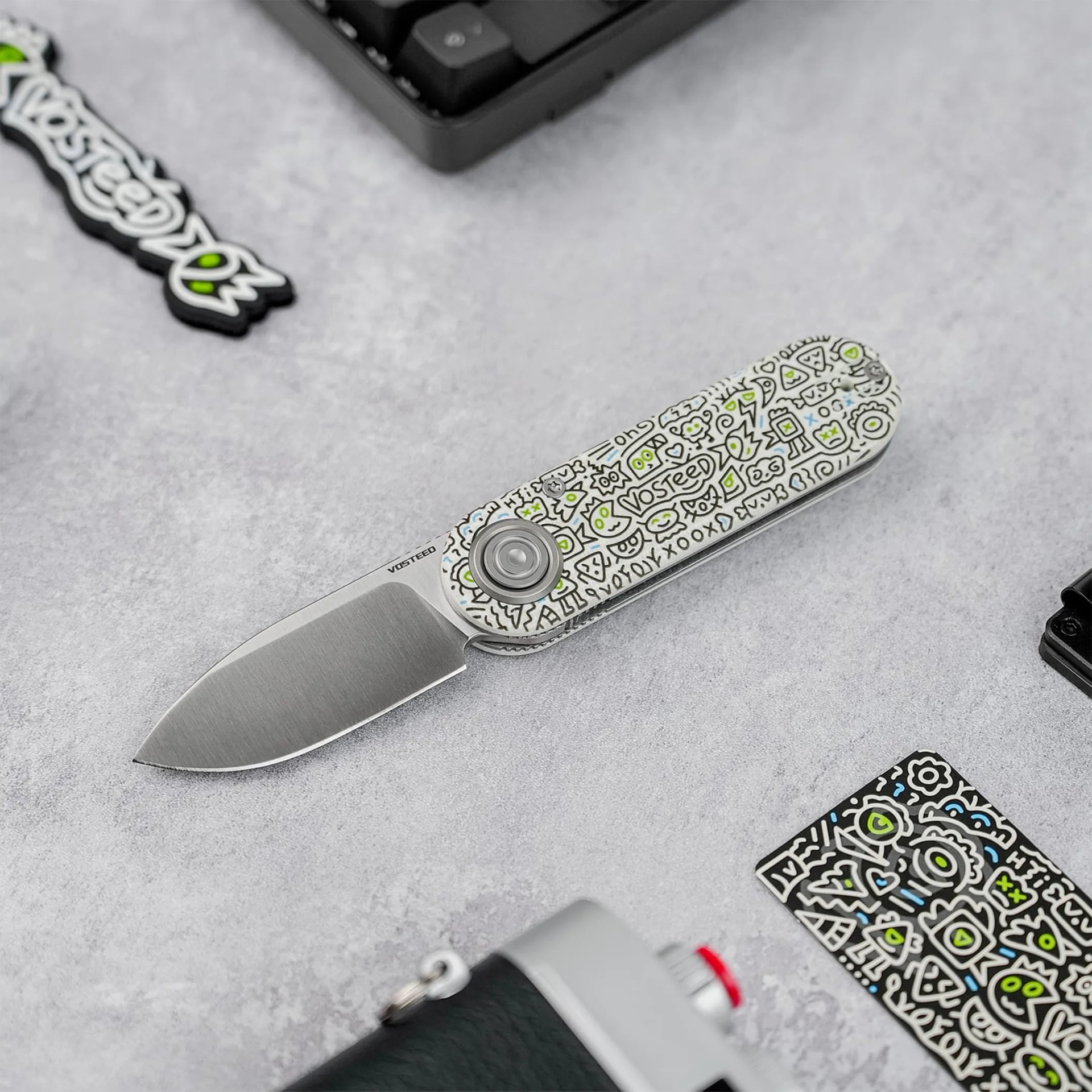 Folding knife with a silver blade and a white handle decorated with black and green doodle patterns, placed on a gray surface.