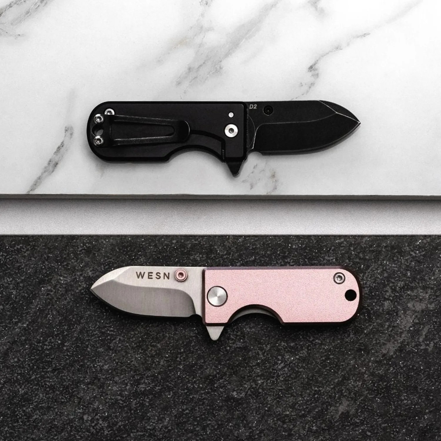 Two small folding knives, one black with a clip and one pink with a silver blade marked "WESN," placed on contrasting marble and dark stone surfaces.