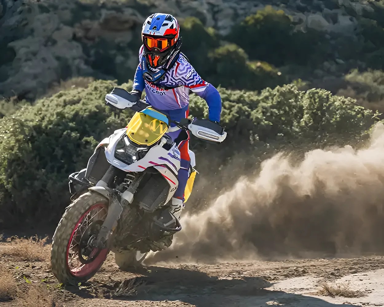 Motorcyclist in blue and white gear riding a white dirt bike with a yellow windshield on a dirt trail kicking up dust.