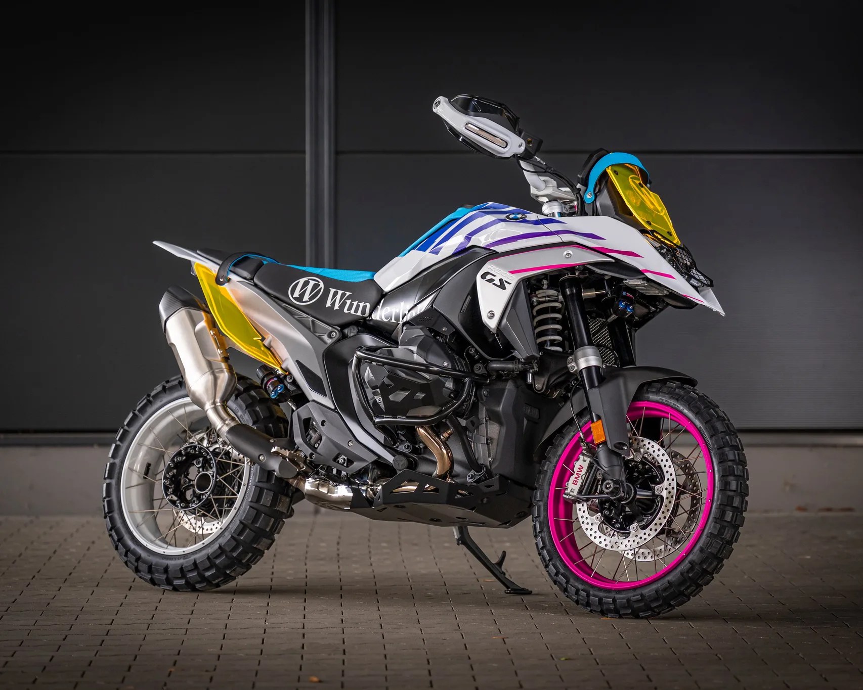White and black BMW GS motorcycle with pink front wheel rim, yellow windscreen, and Wunderlich branding on the seat.