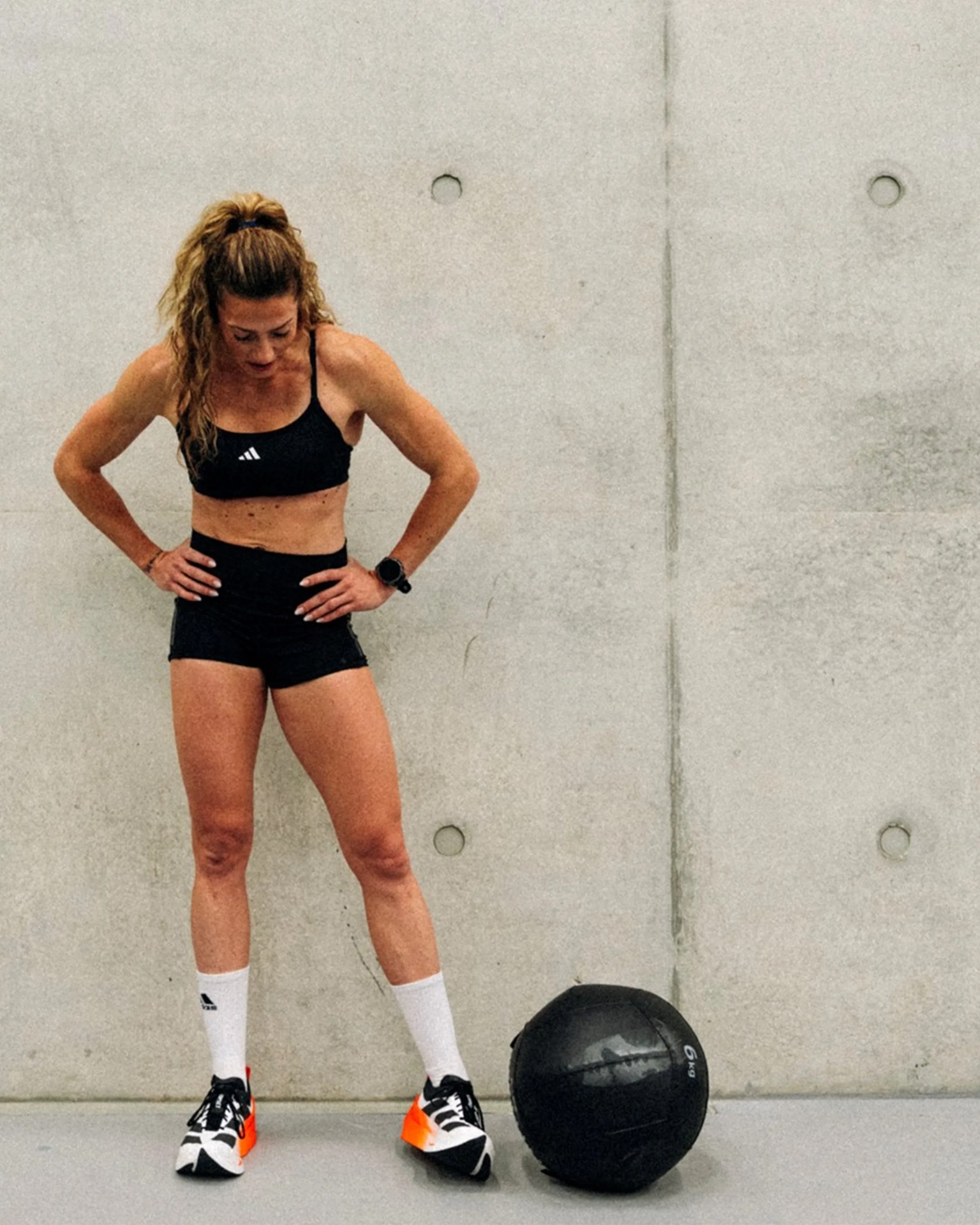 Woman in black sports bra and shorts with white socks and black-and-orange sneakers standing next to a black 6 kg medicine ball against a concrete wall.