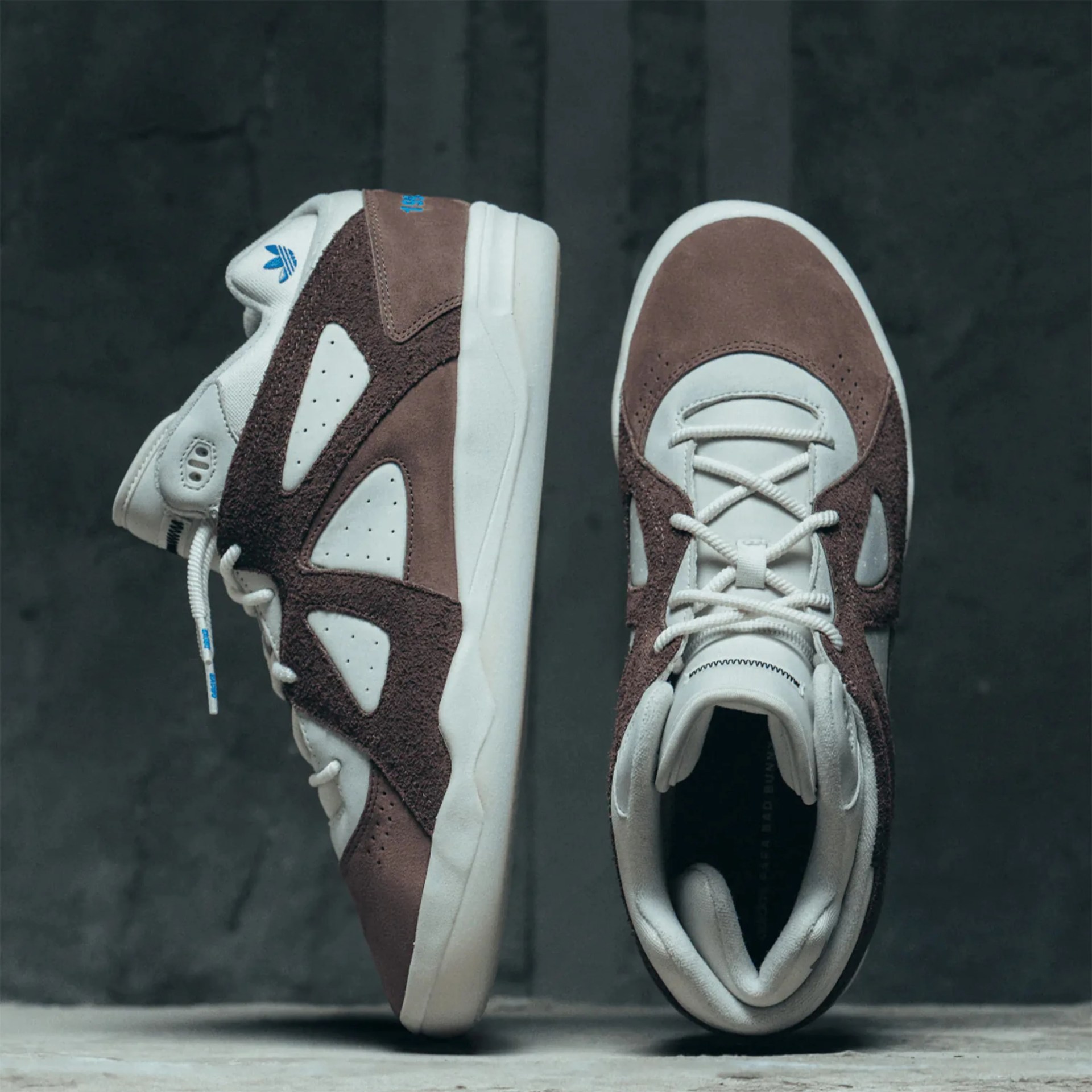 Pair of white and brown suede Adidas sneakers with blue logo accents on a gray surface.