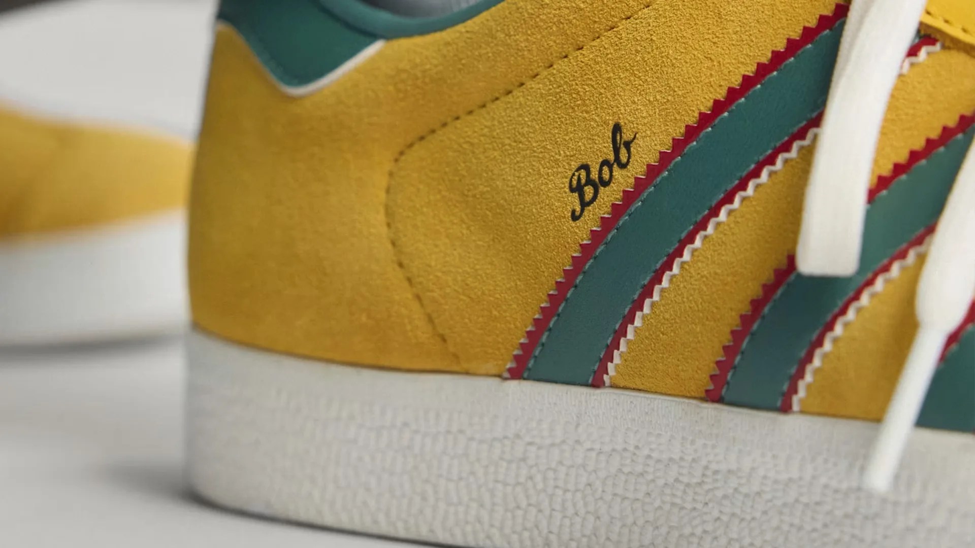 Close-up of a yellow suede sneaker with teal and red zigzag stripes and white laces, featuring the name "Bob.