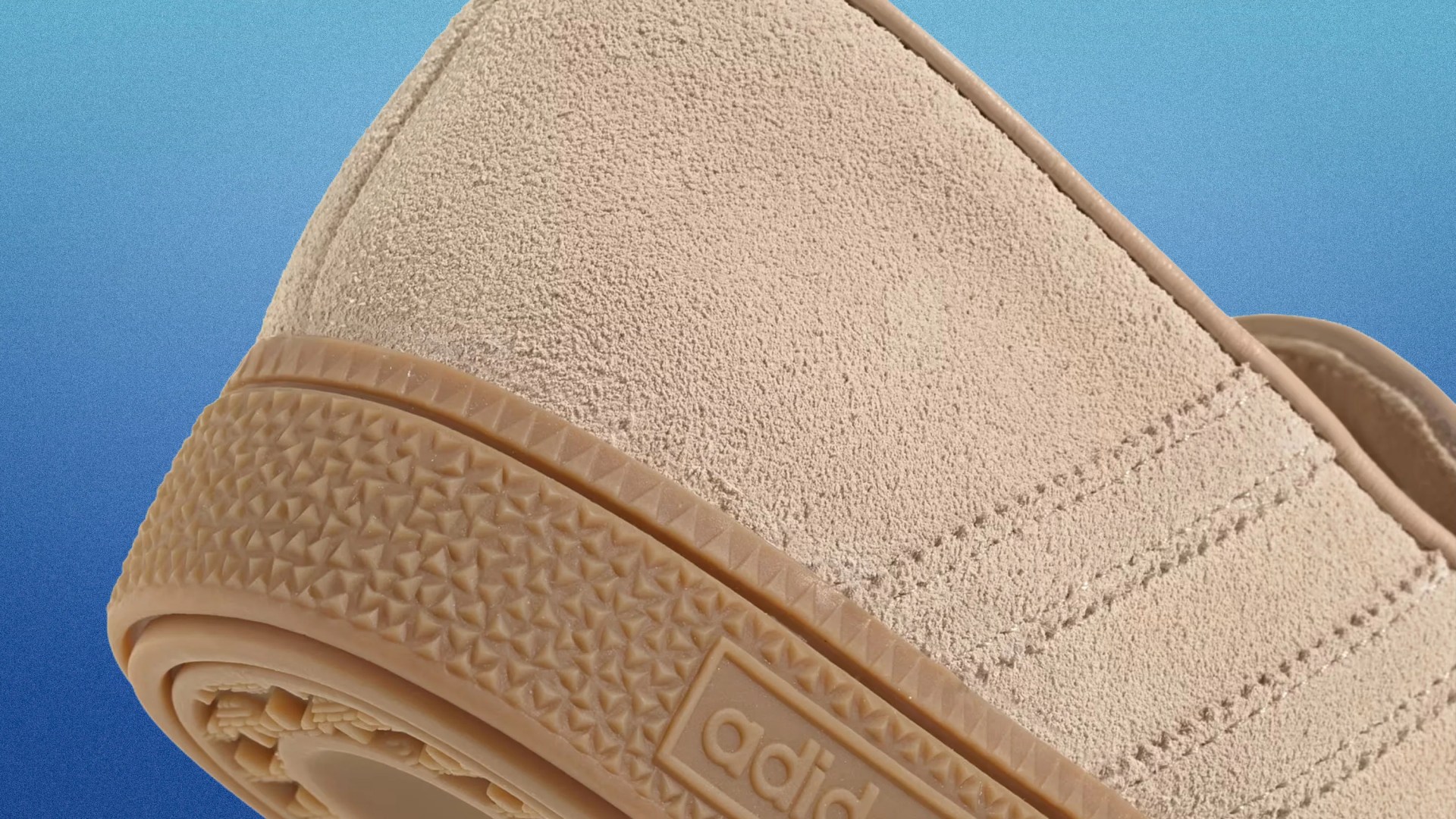 Close-up of beige suede Adidas shoe heel with textured gum sole and partial logo on a blue gradient background.