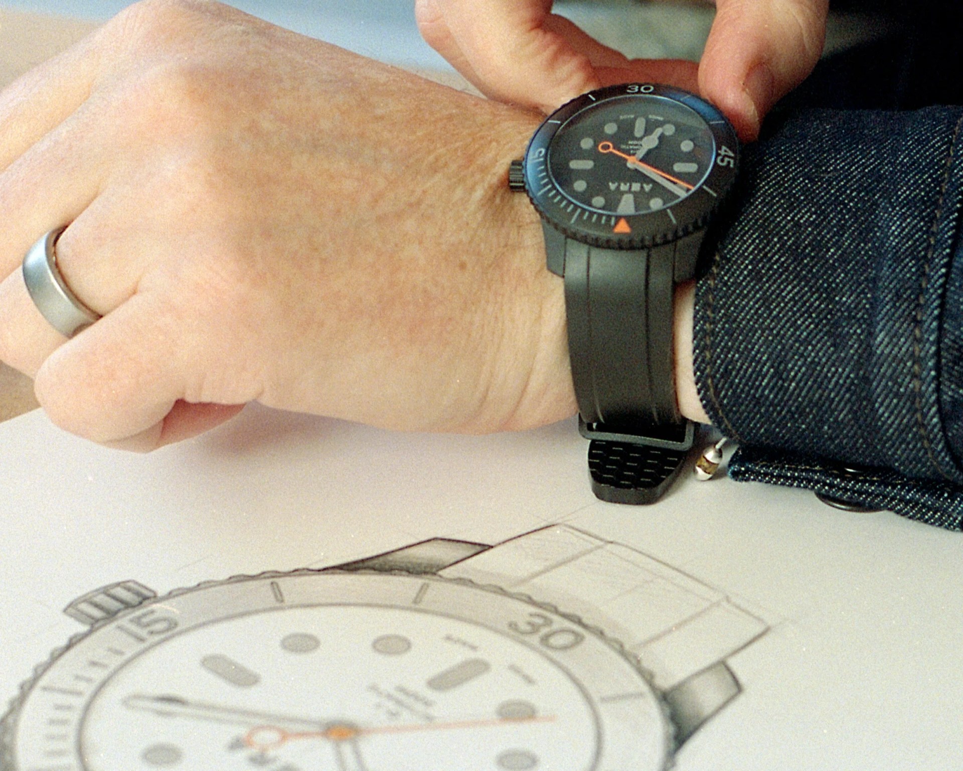 Black dive watch with orange second hand worn on a wrist above a pencil sketch of the same watch.