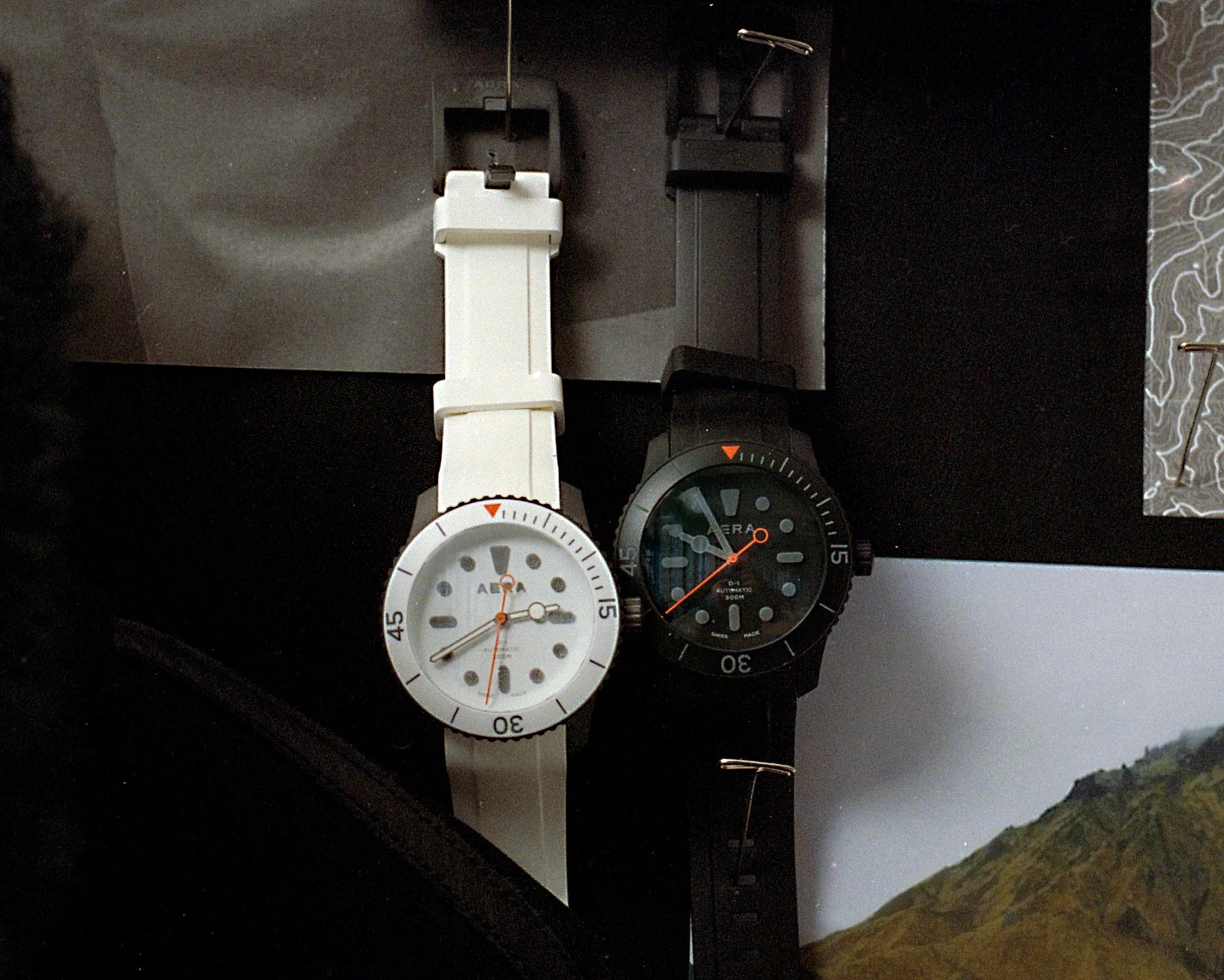 Two wristwatches, one white and one black, with round faces and rubber straps, placed side by side on a dark surface.