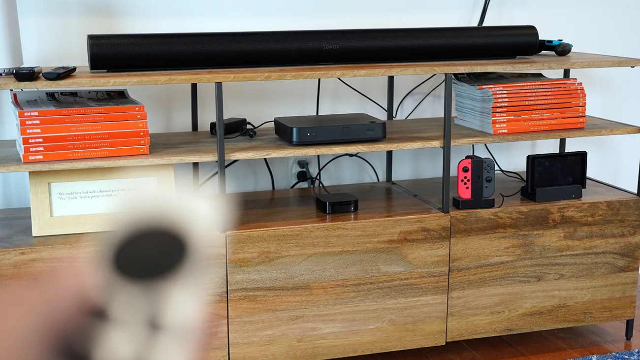 Wooden entertainment shelf with black Sonos soundbar, stacks of orange books, black media devices, and a Nintendo Switch console.
