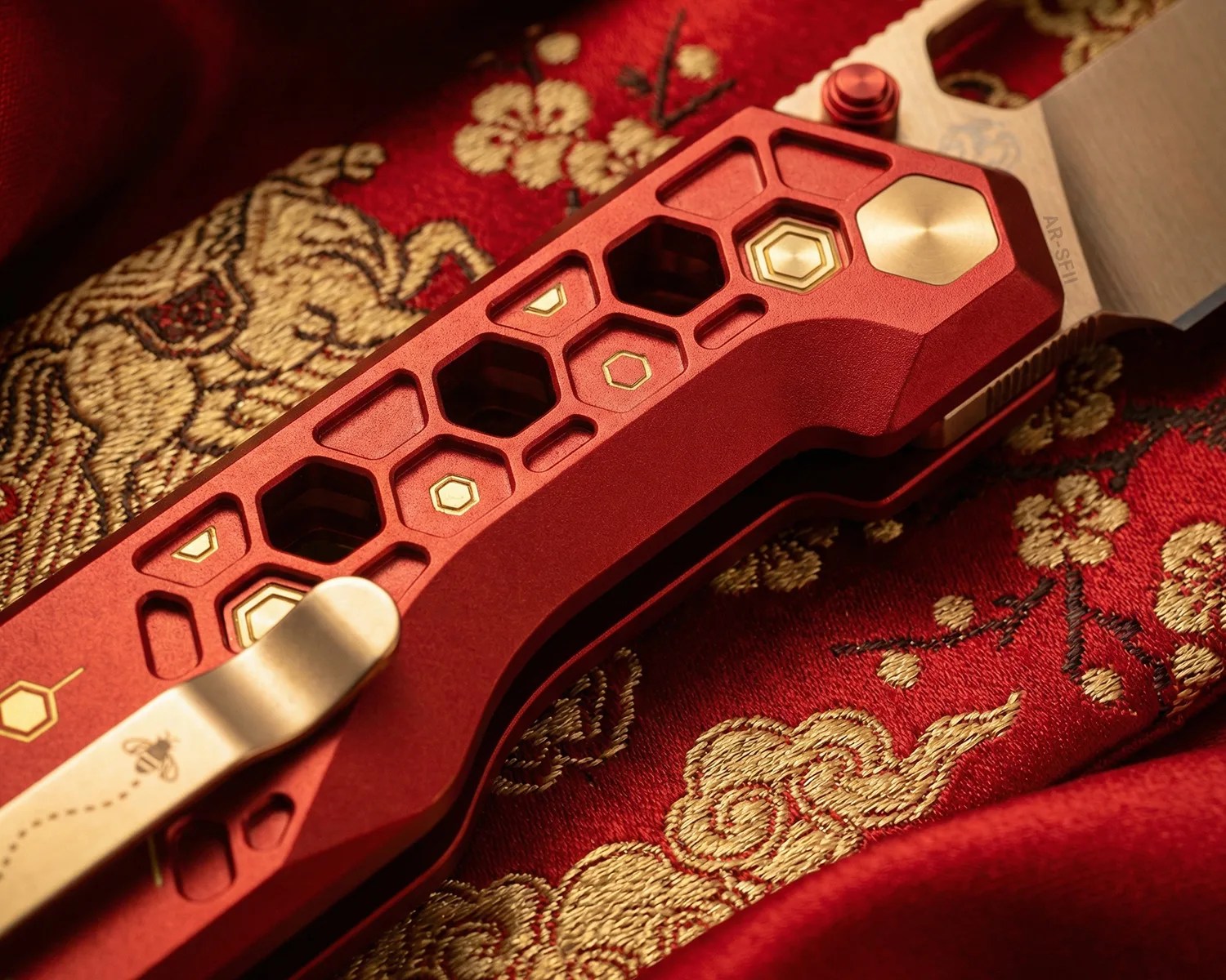 Close-up of a red folding knife handle with hexagonal cutouts and gold accents resting on red and gold embroidered fabric.