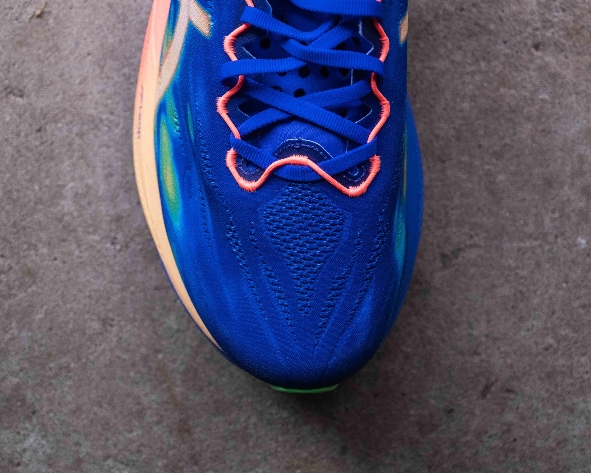 Close-up of a blue sneaker toe with orange lace loops and a beige sole on a concrete surface.