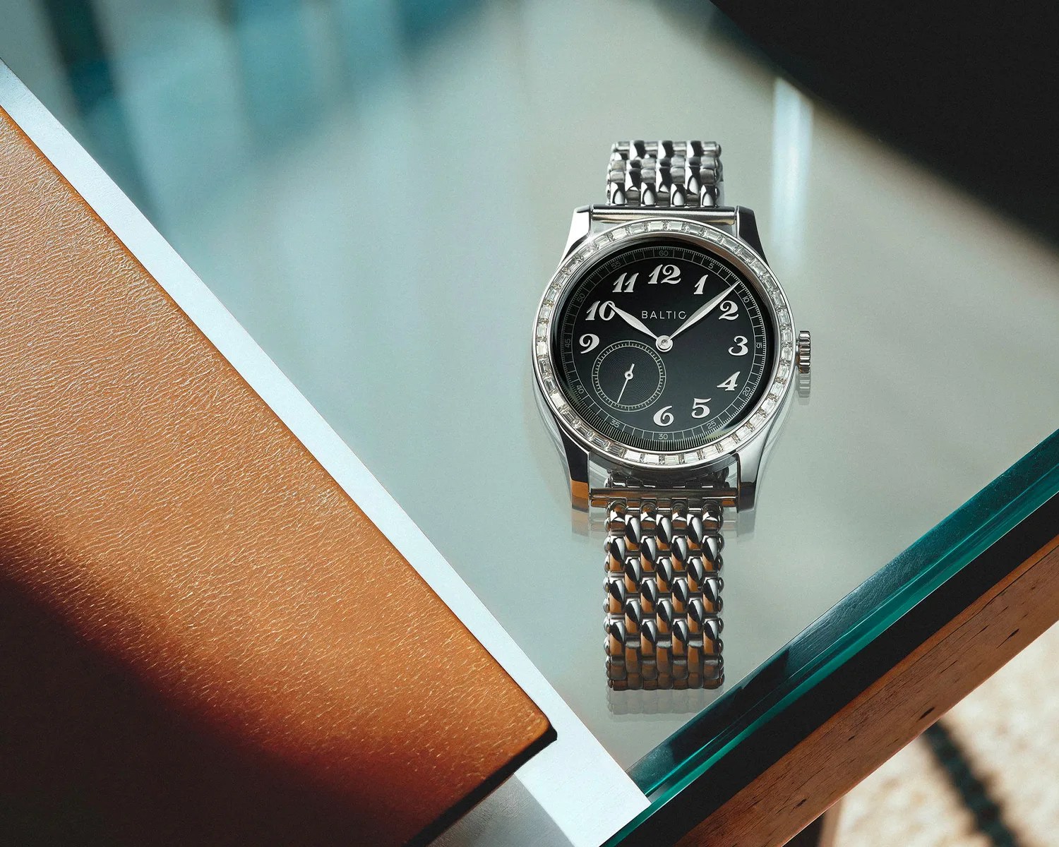 Silver Baltic wristwatch with black dial and Arabic numerals on a glass table next to a brown leather notebook.