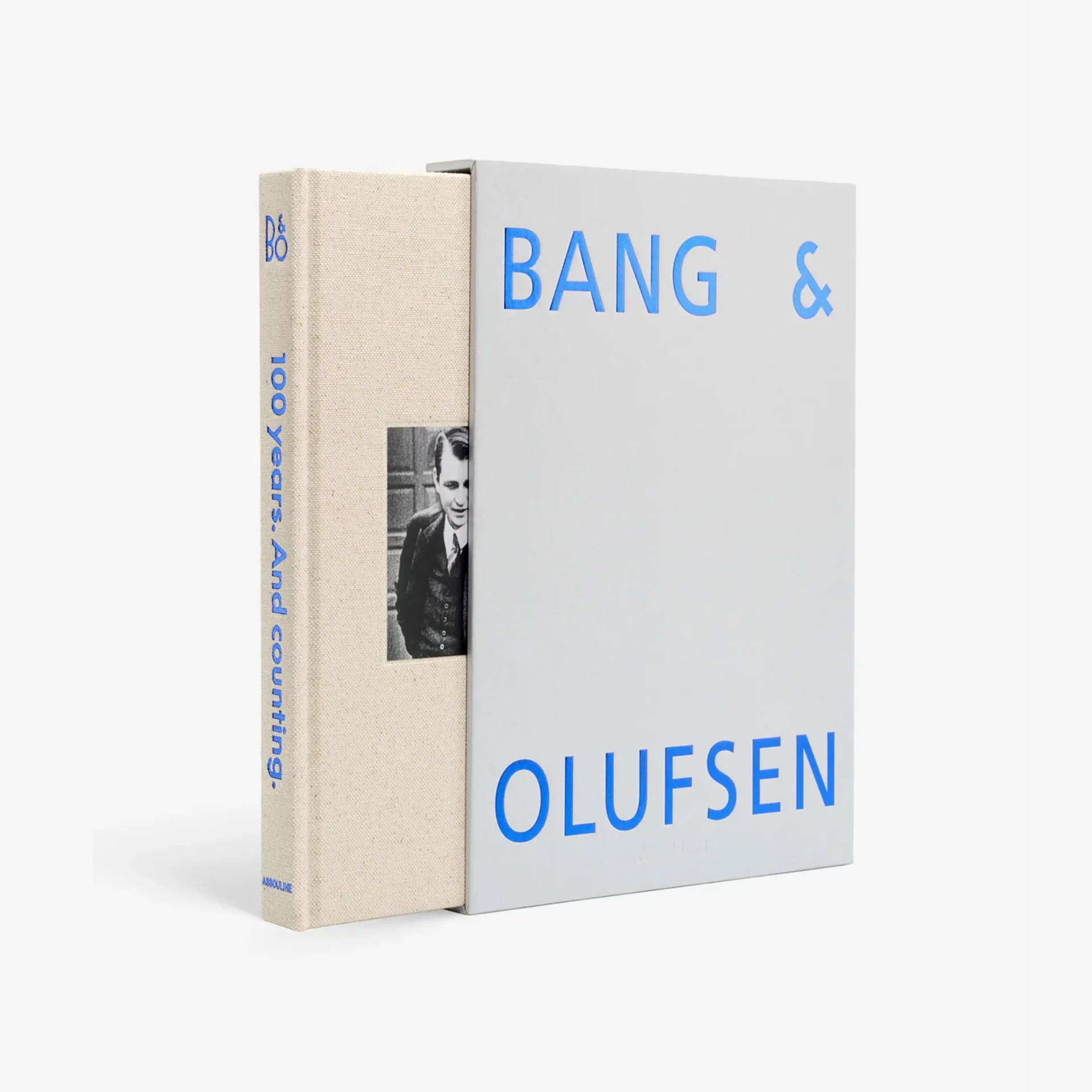 Beige cloth-bound book titled "100 Years. And counting." with a black-and-white photo on the spine, inside a gray slipcase with blue "BANG & OLUFSEN" text.