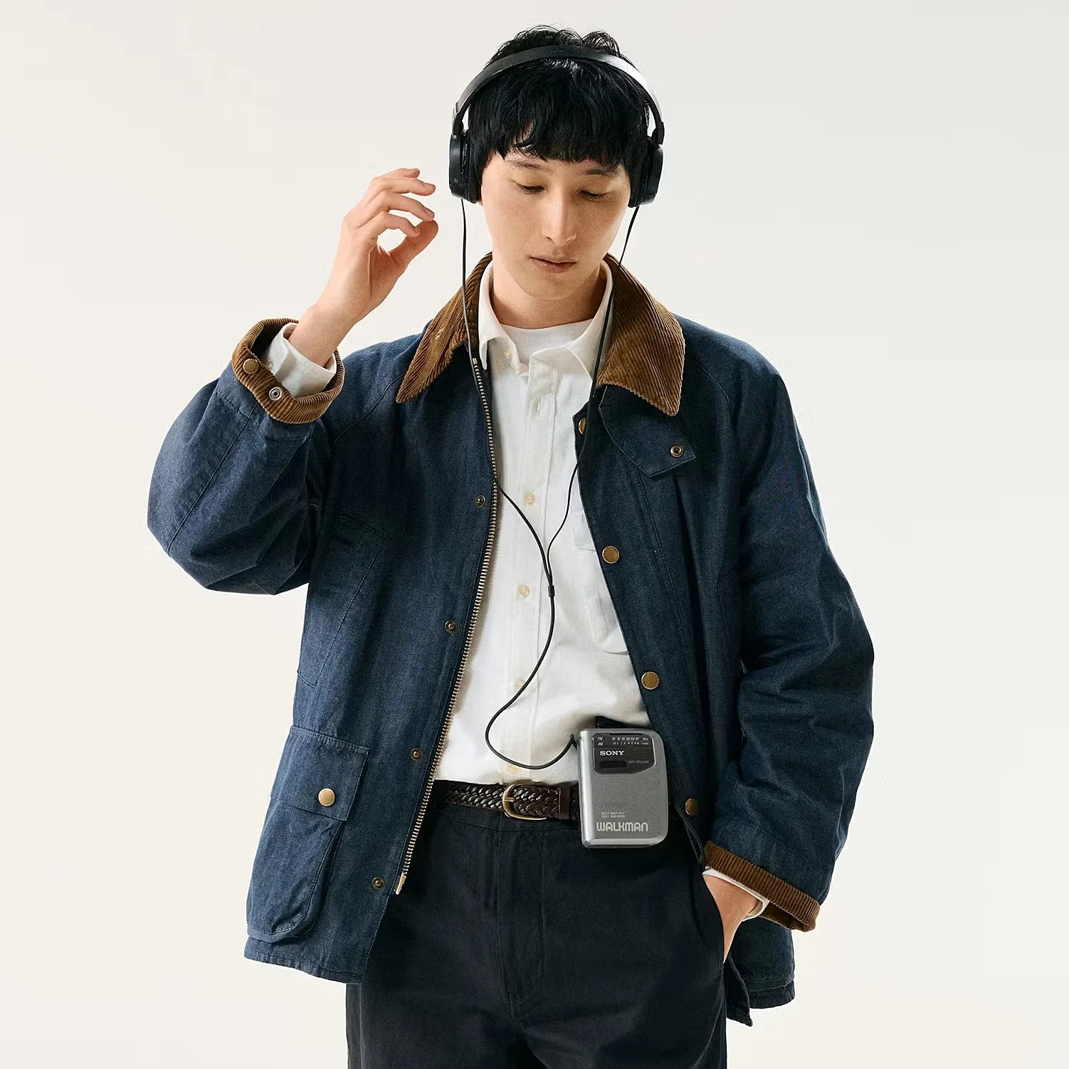 Person wearing a dark blue jacket with brown corduroy collar and cuffs, white shirt, black pants, and black headphones connected to a silver Sony Walkman clipped to their waist.