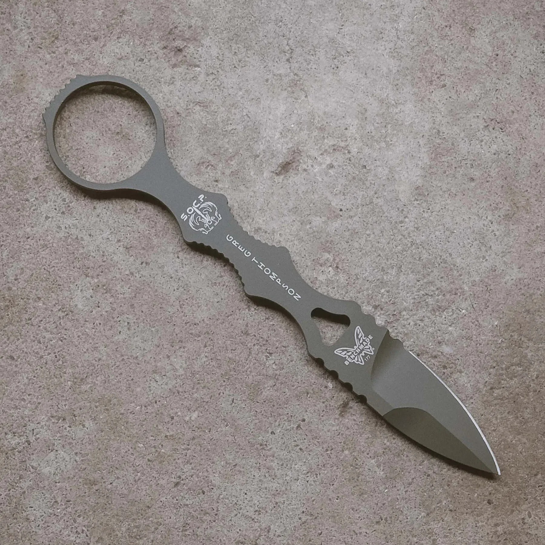 Gray tactical knife with a ring handle and serrated edges on a textured surface.