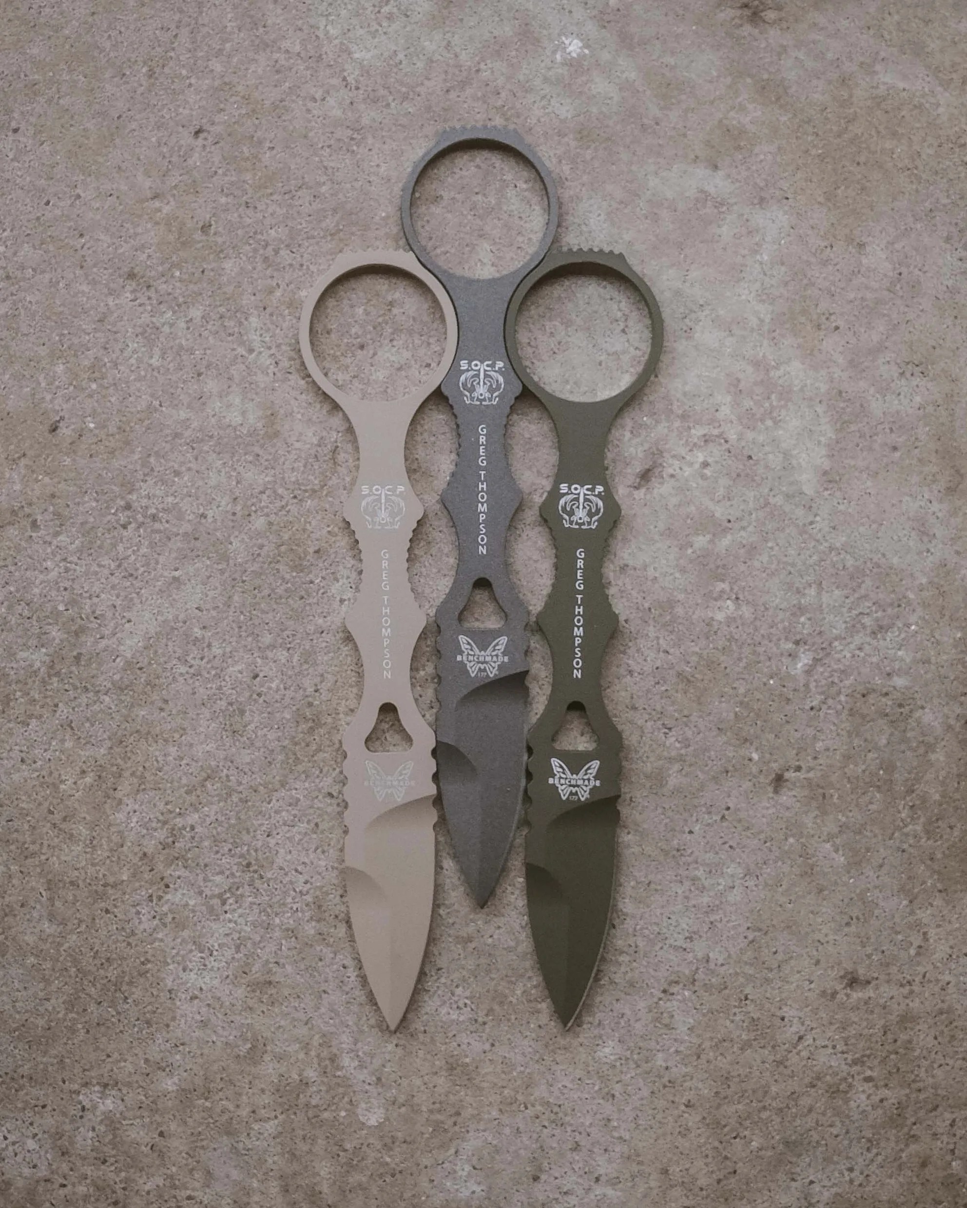 Three tactical push daggers in tan, gray, and olive green with finger rings and engraved logos on a textured surface.
