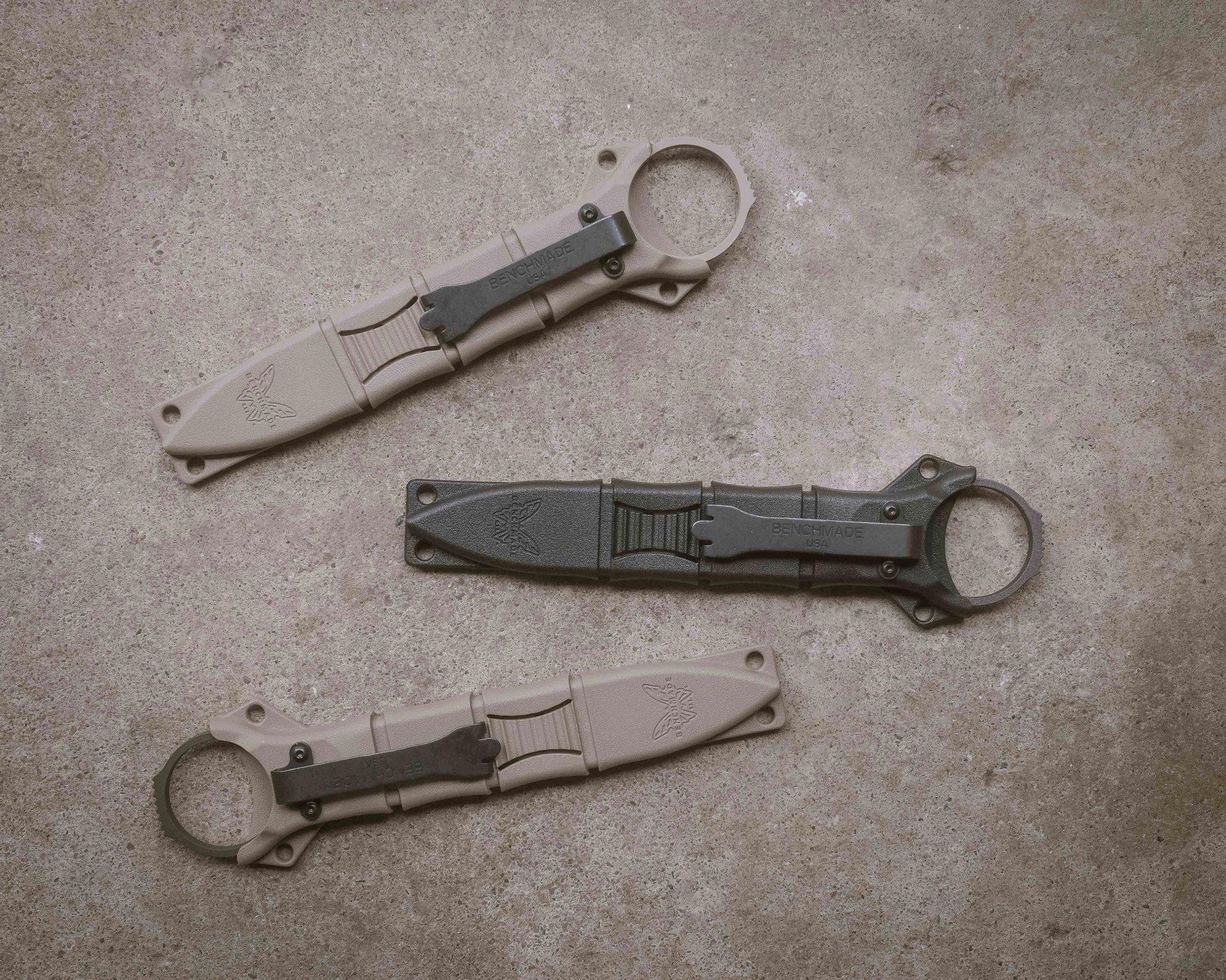 Three Benchmade knives with sheaths, two tan and one black, laid out on a textured concrete surface.