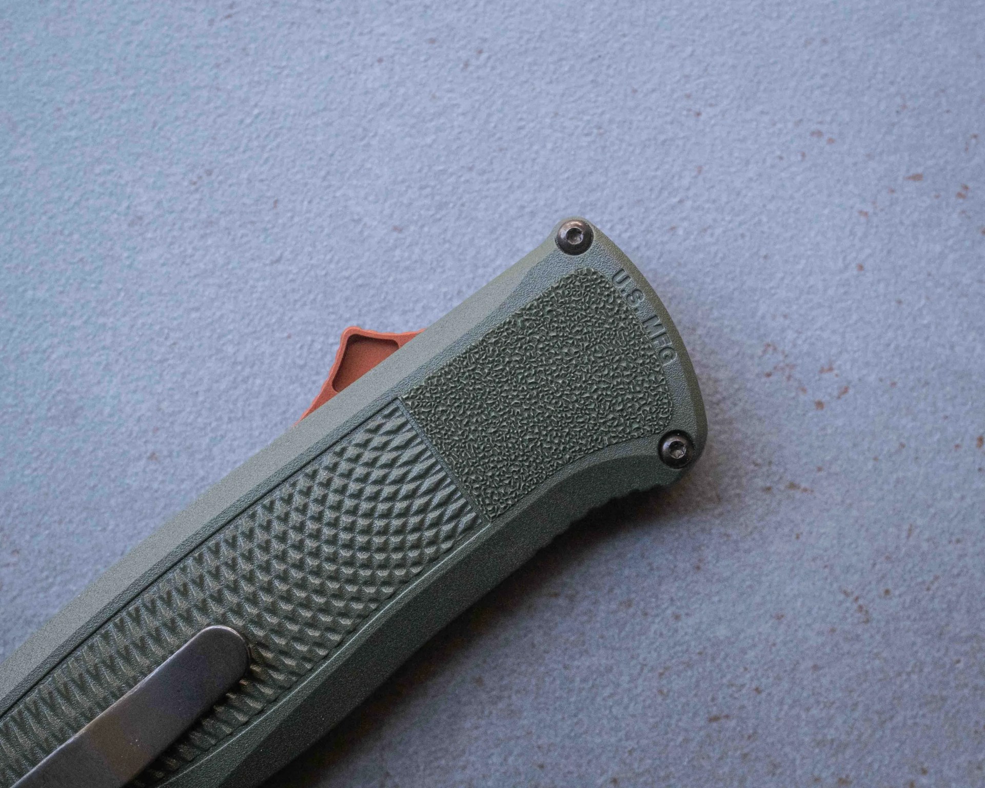 Close-up of textured green knife handle with "U.S. MFG" engraved and a black metal clip on a gray surface.