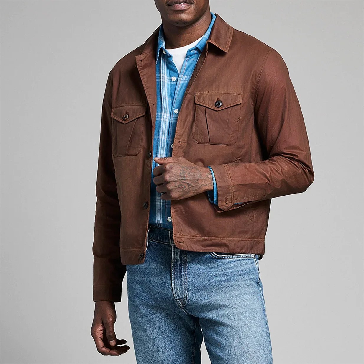 Brown button-up jacket with flap chest pockets worn over a blue plaid shirt and light blue jeans.