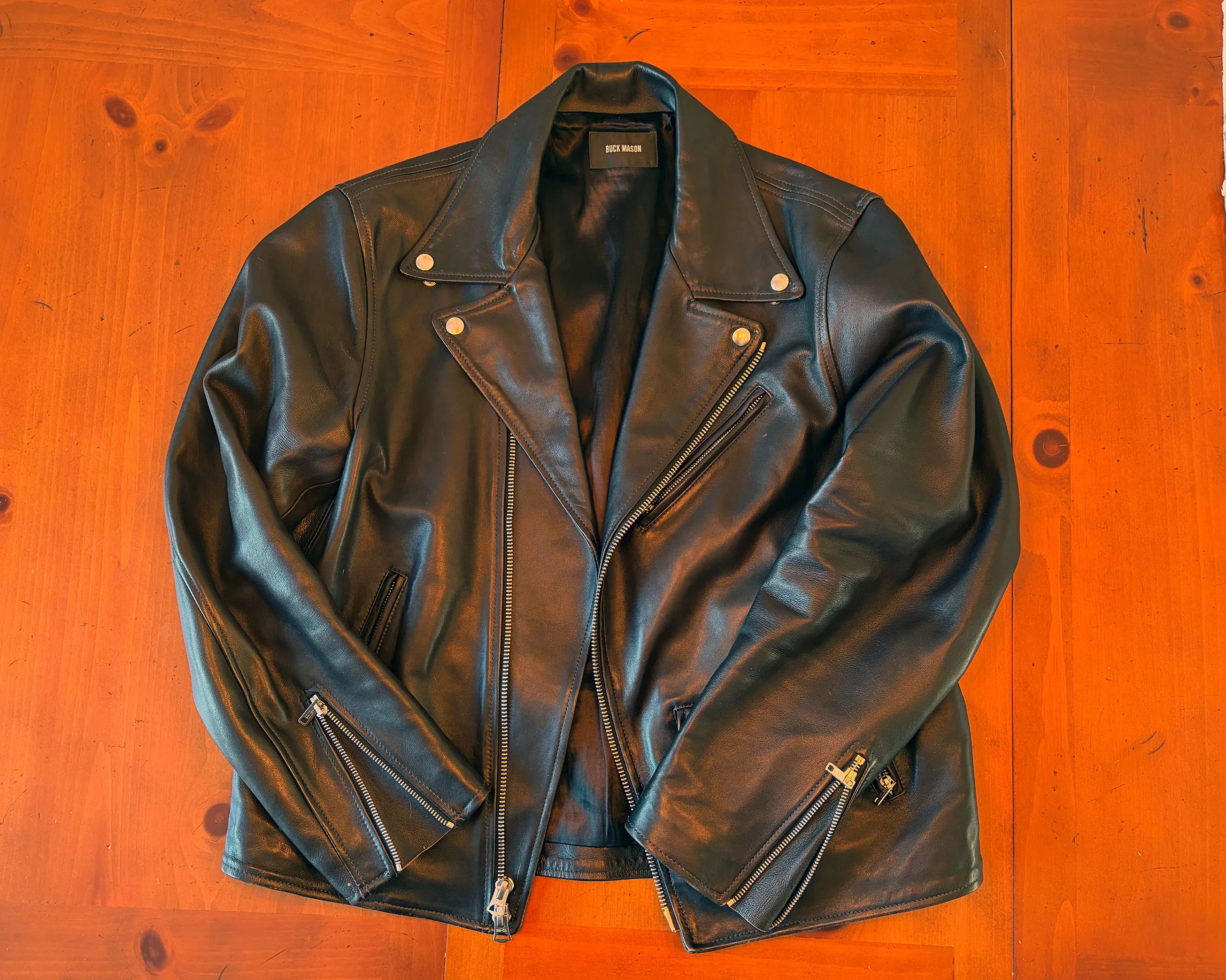 Black leather biker jacket with silver zippers and snap buttons laid flat on a wooden surface.