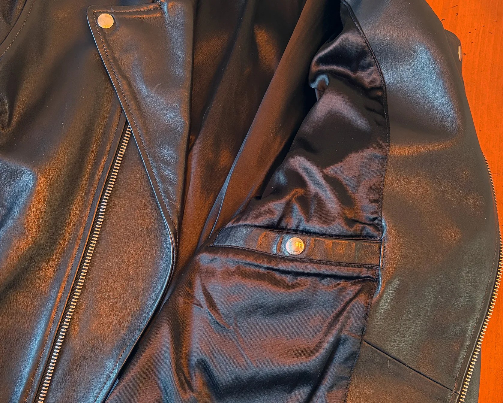 Black leather jacket with silver zipper and snap button pocket on a wooden surface.