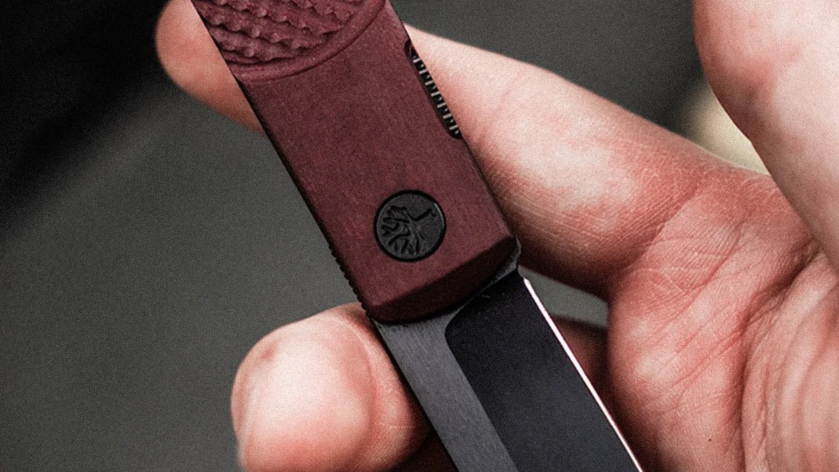 Hand holding a folding knife with a dark red textured handle and a black blade.