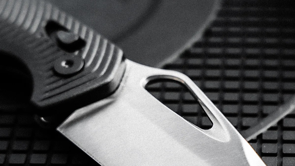 Close-up of a folding knife blade with a thumb hole and textured black handle on a grid surface.