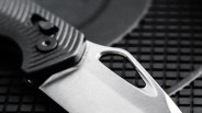 Close-up of a folding knife blade with a thumb hole and textured black handle on a grid surface.