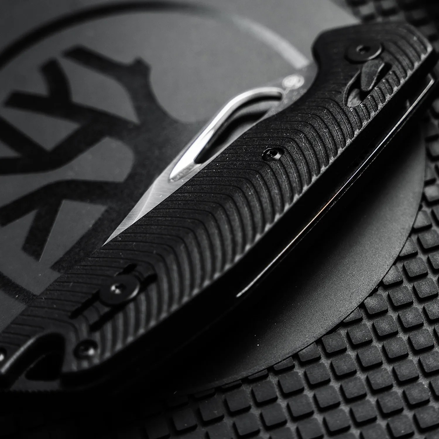 Black folding knife with textured handle and partially visible silver blade on a patterned black surface.