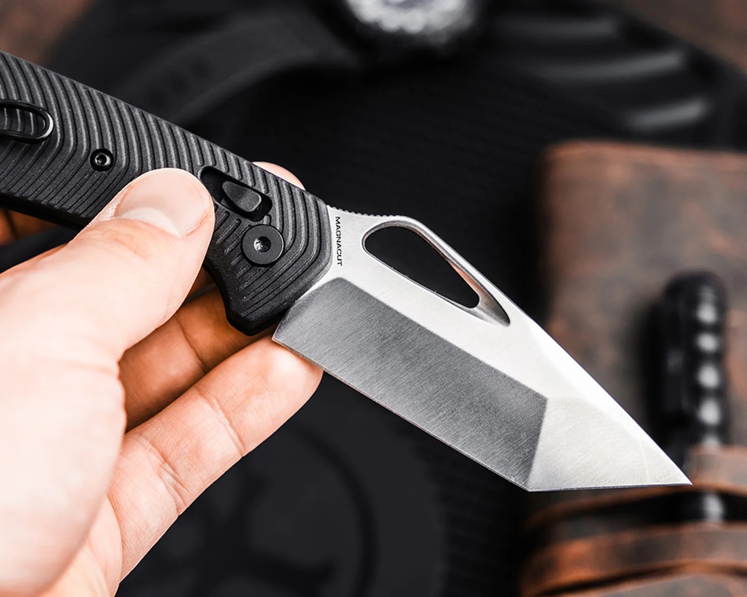 Hand holding a folding knife with a black textured handle and a silver tanto-style blade marked "MAGNACUT.