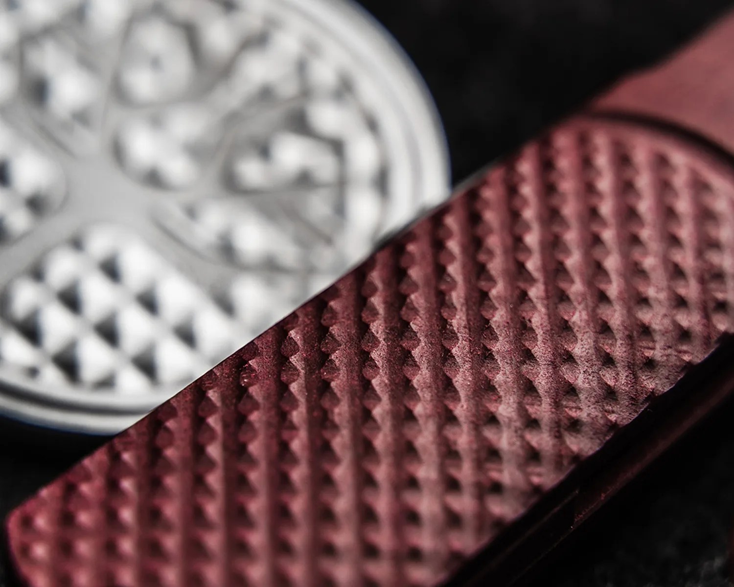 Close-up of a textured maroon metal surface with a blurred silver circular object in the background.