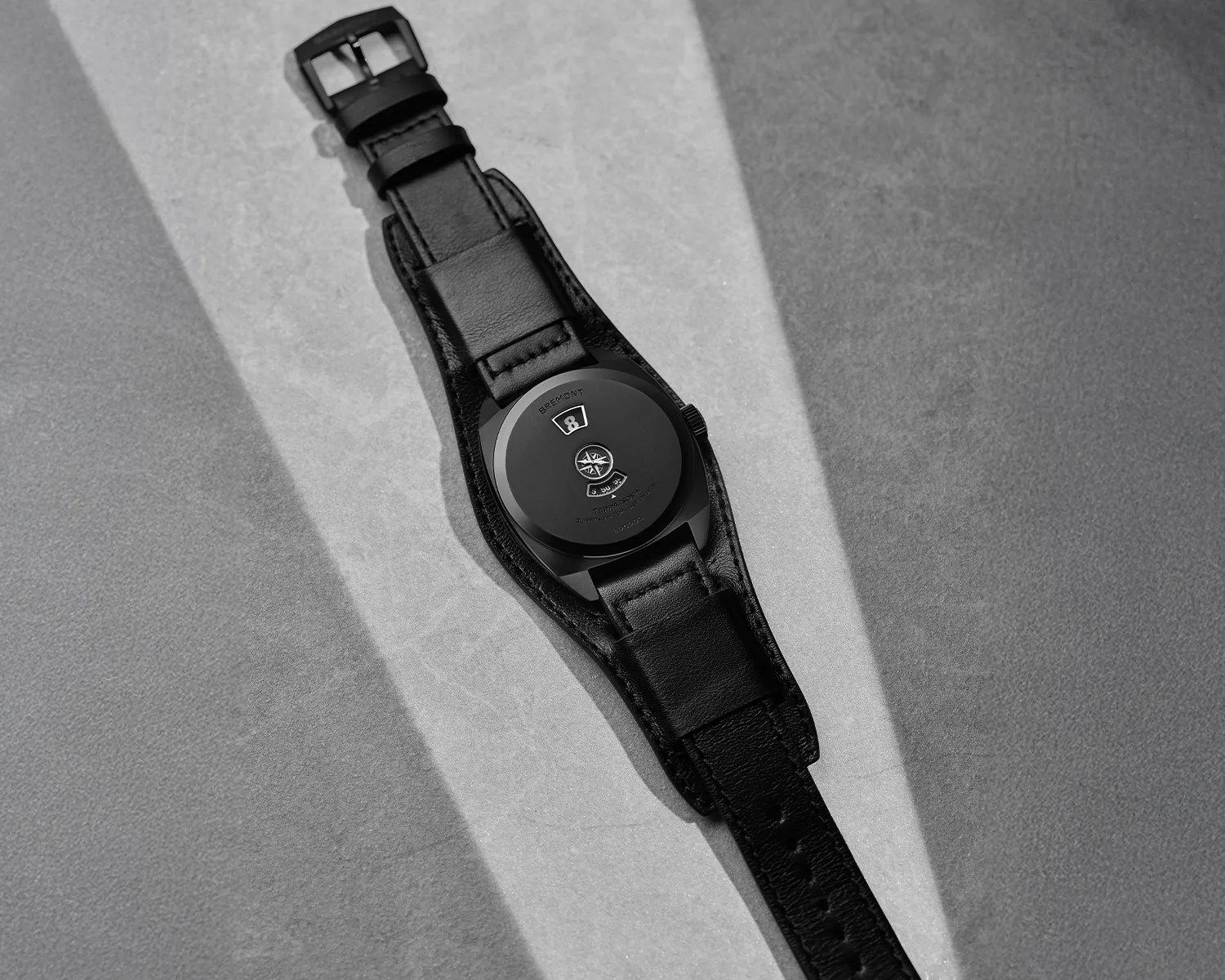Black leather wristwatch with a black case and unique back design, placed on a textured surface with light and shadow.