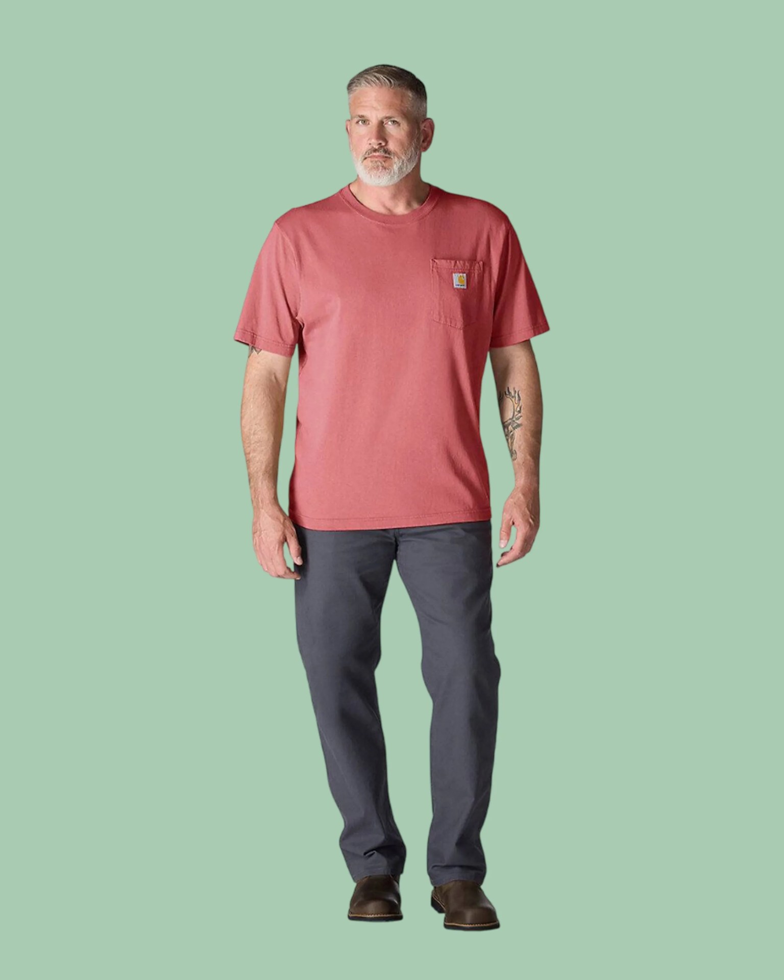 Man wearing a faded red short-sleeve pocket t-shirt, dark gray pants, and brown shoes against a green background.