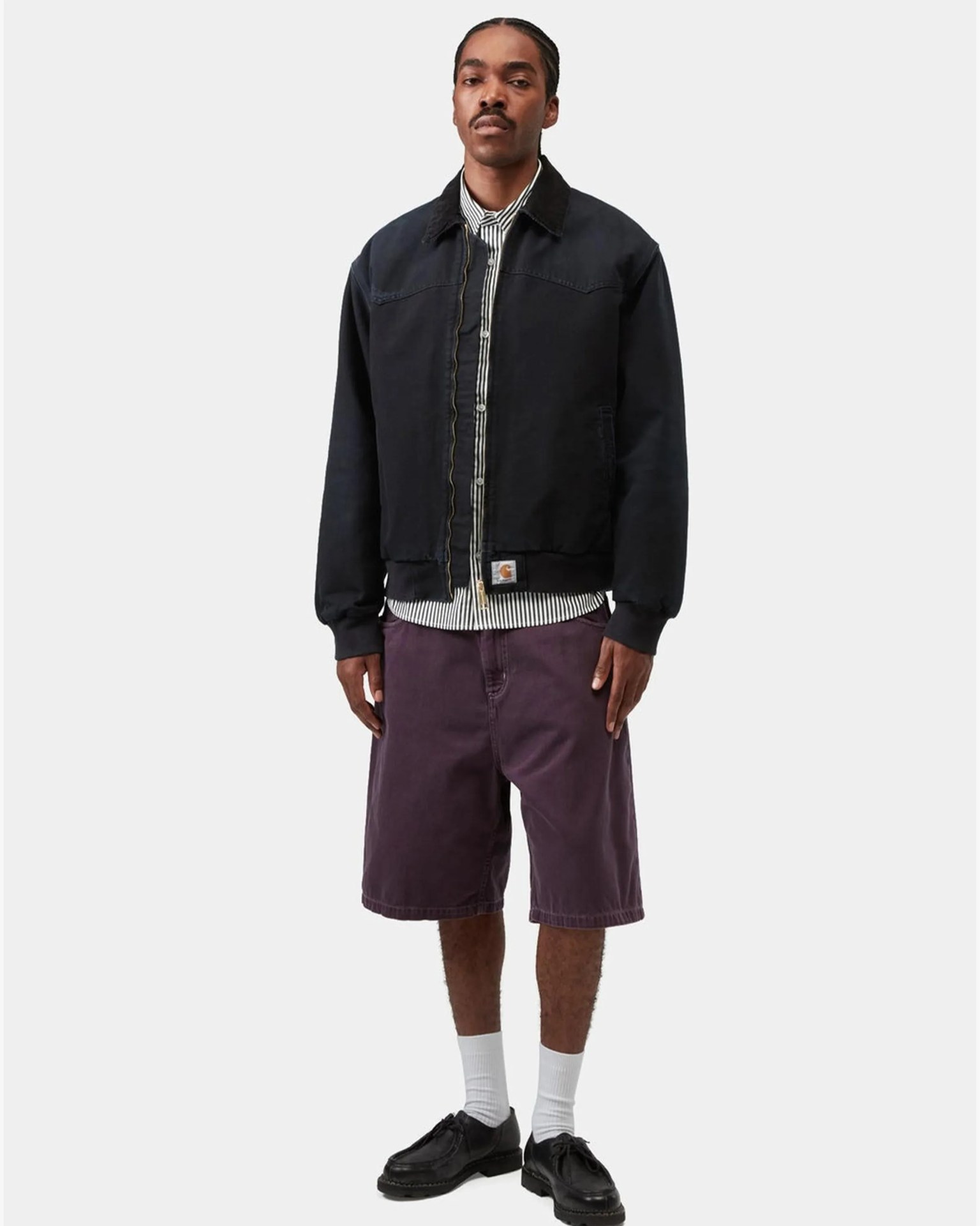 Man wearing a black zip-up jacket over a black and white striped shirt, purple knee-length shorts, white socks, and black shoes.