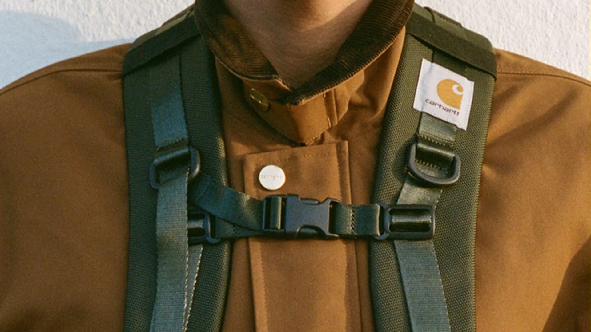 Close-up of green backpack straps with a black buckle worn over a brown Carhartt jacket.