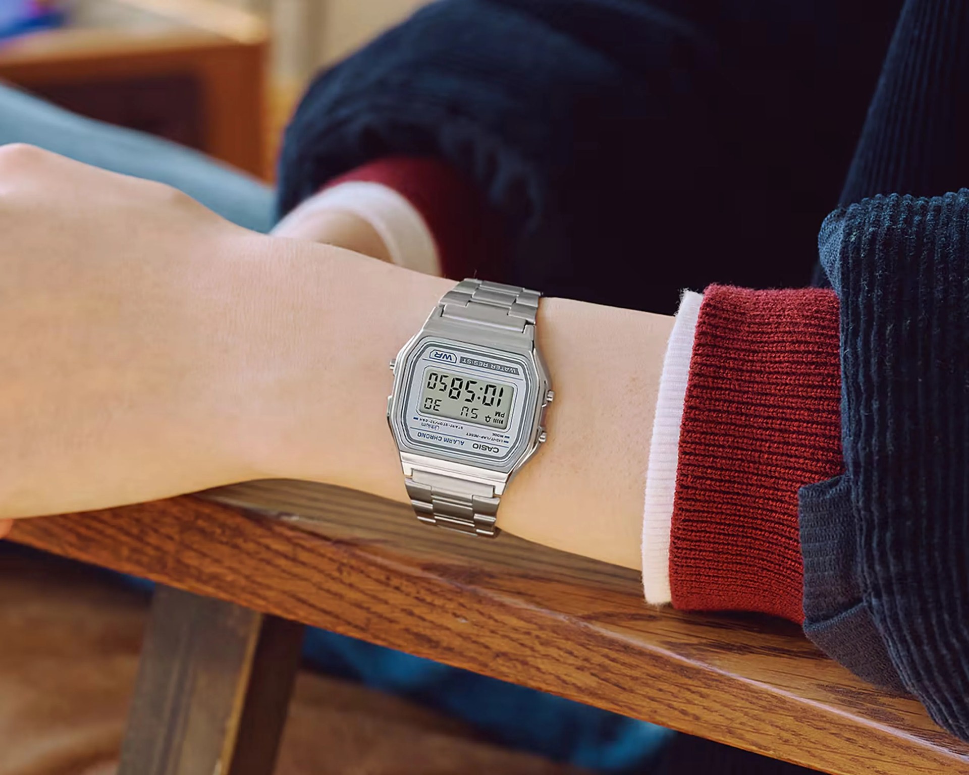 Silver Casio digital watch with metal band worn on a wrist resting on a wooden surface.
