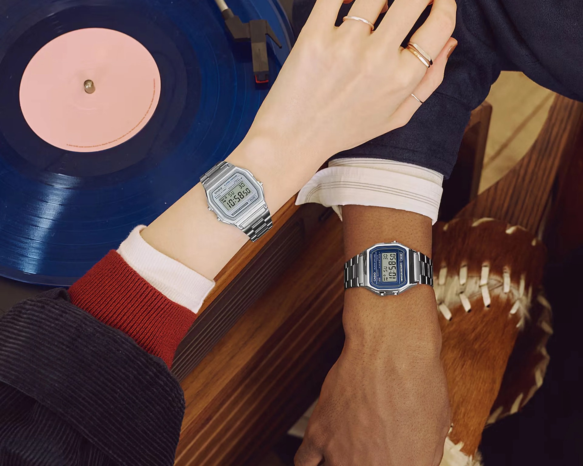 Two wrists wearing silver digital Casio watches, one with a blue face and the other with a silver face, near a blue vinyl record player.