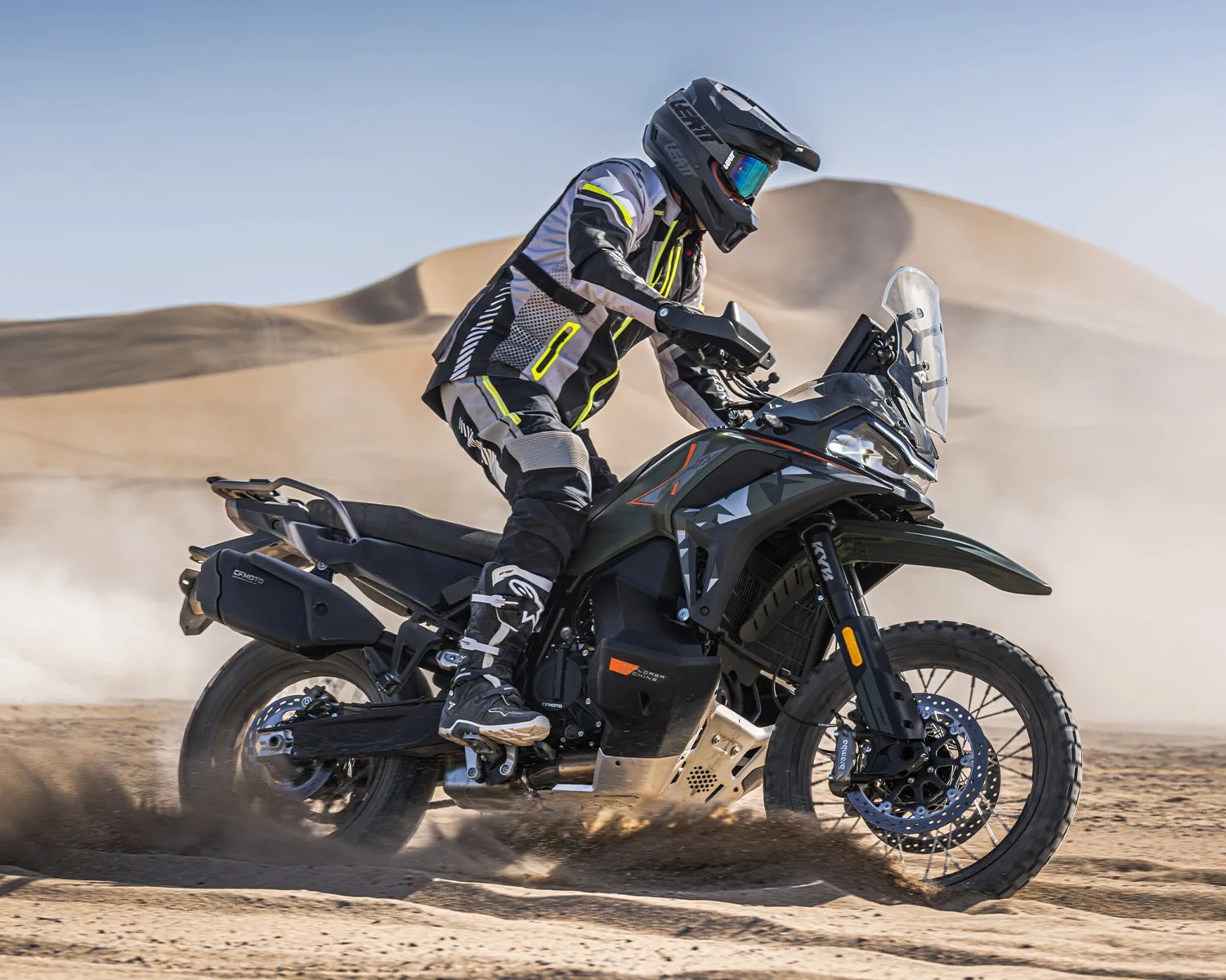 Person wearing black and gray riding gear and helmet on a black adventure motorcycle kicking up sand in a desert.