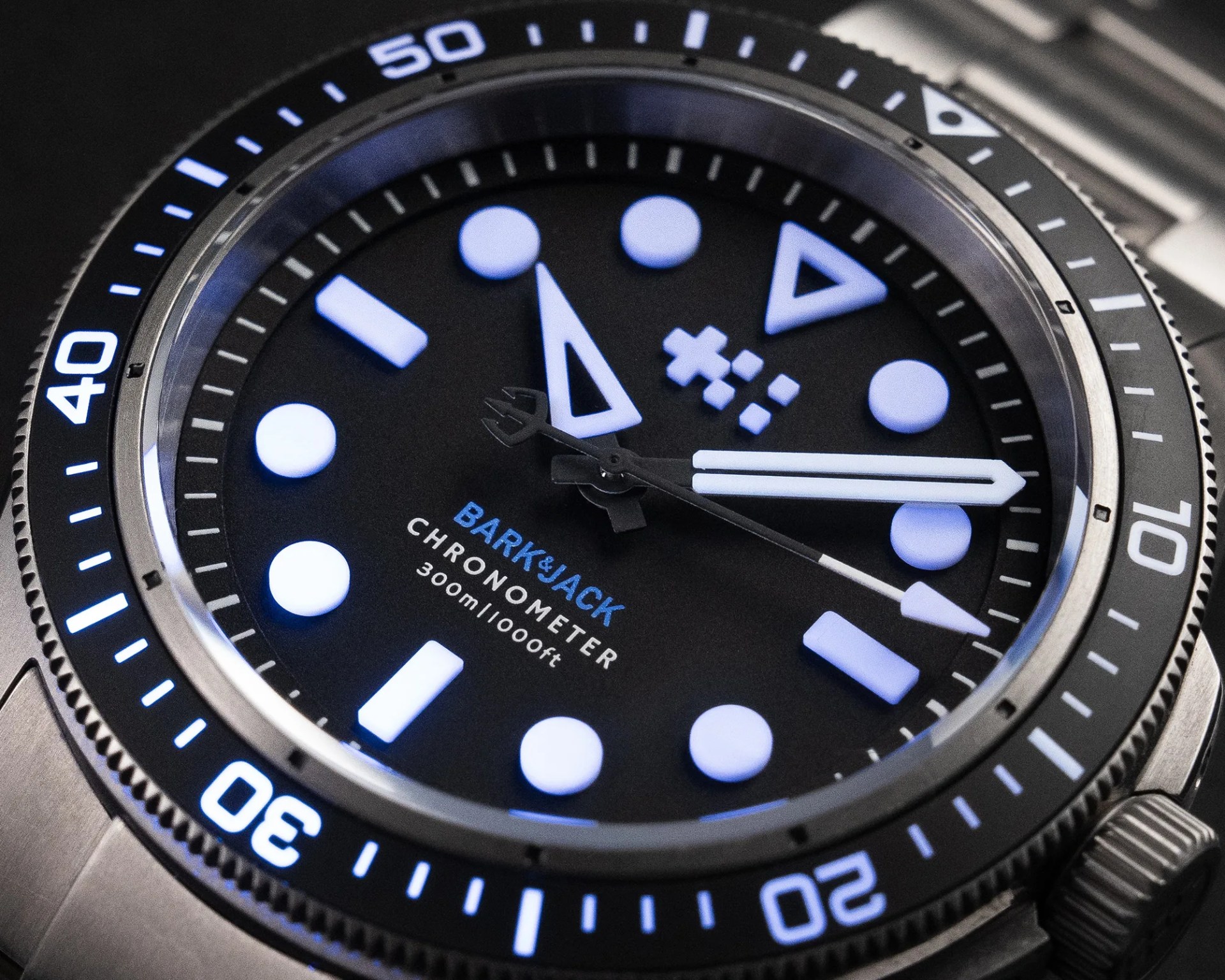 Close-up of a Bark & Jack chronometer watch face with luminous white markers and hands on a black dial and stainless steel bezel.