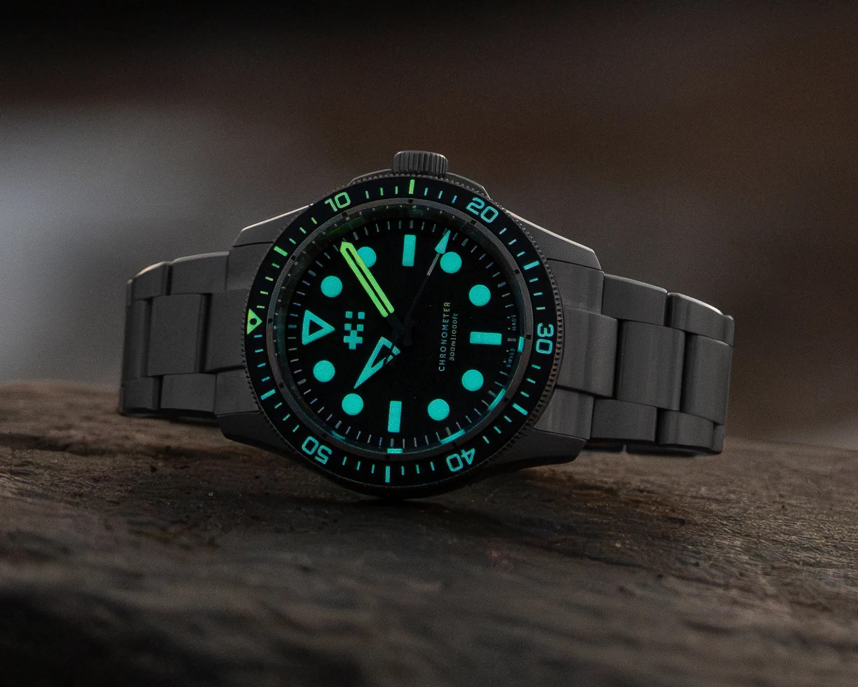 Black metal wristwatch with glowing blue-green hour markers and hands on a dark textured surface.