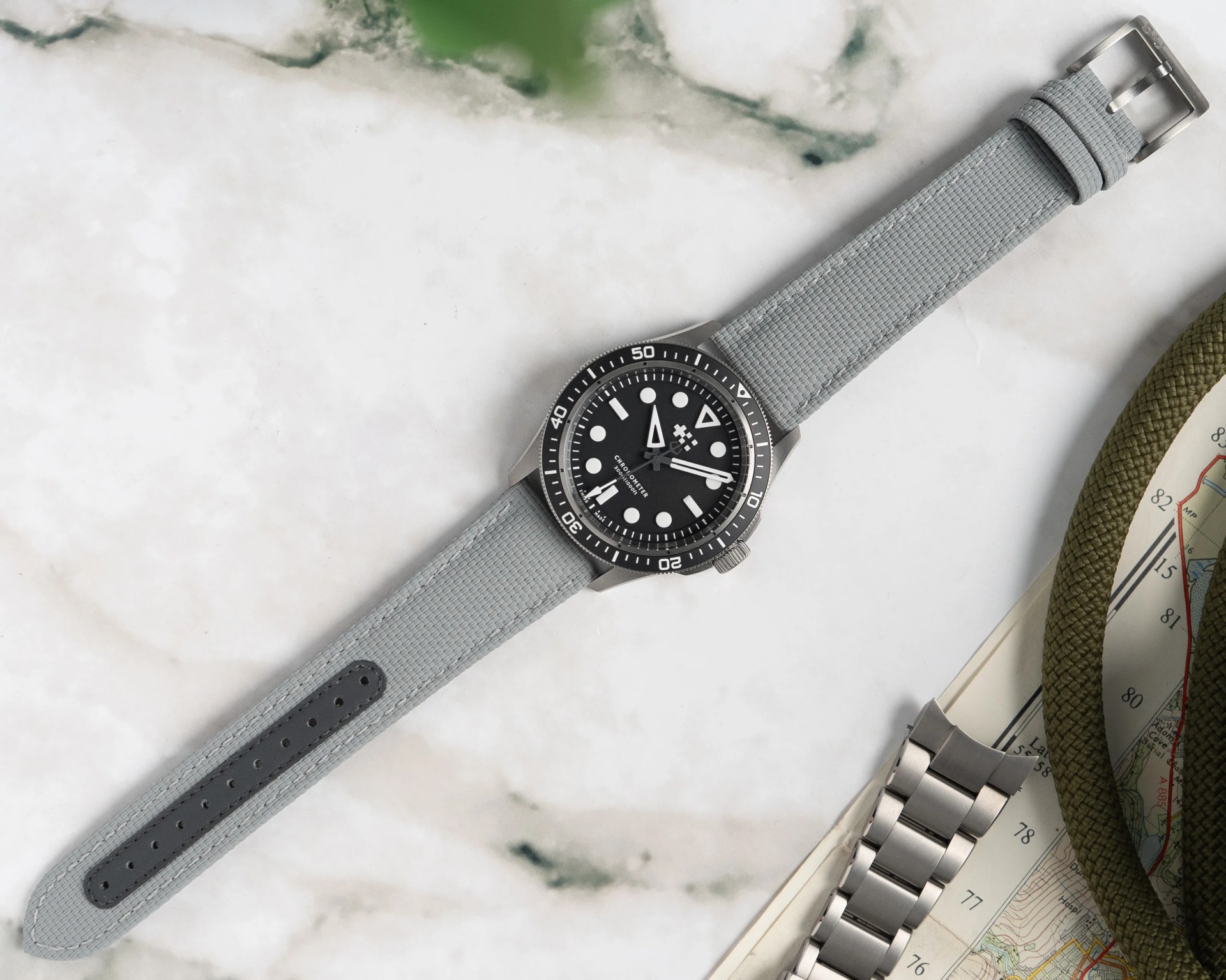 Black dial wristwatch with white markers and a gray fabric strap on a marble surface next to a metal watch band and a map.