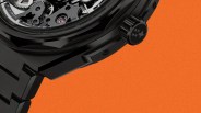 Close-up of a black metal watch crown and part of the bracelet against an orange background.