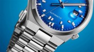 Close-up of a silver metal wristwatch with a blue dial, automatic movement, and date window showing the number 7.