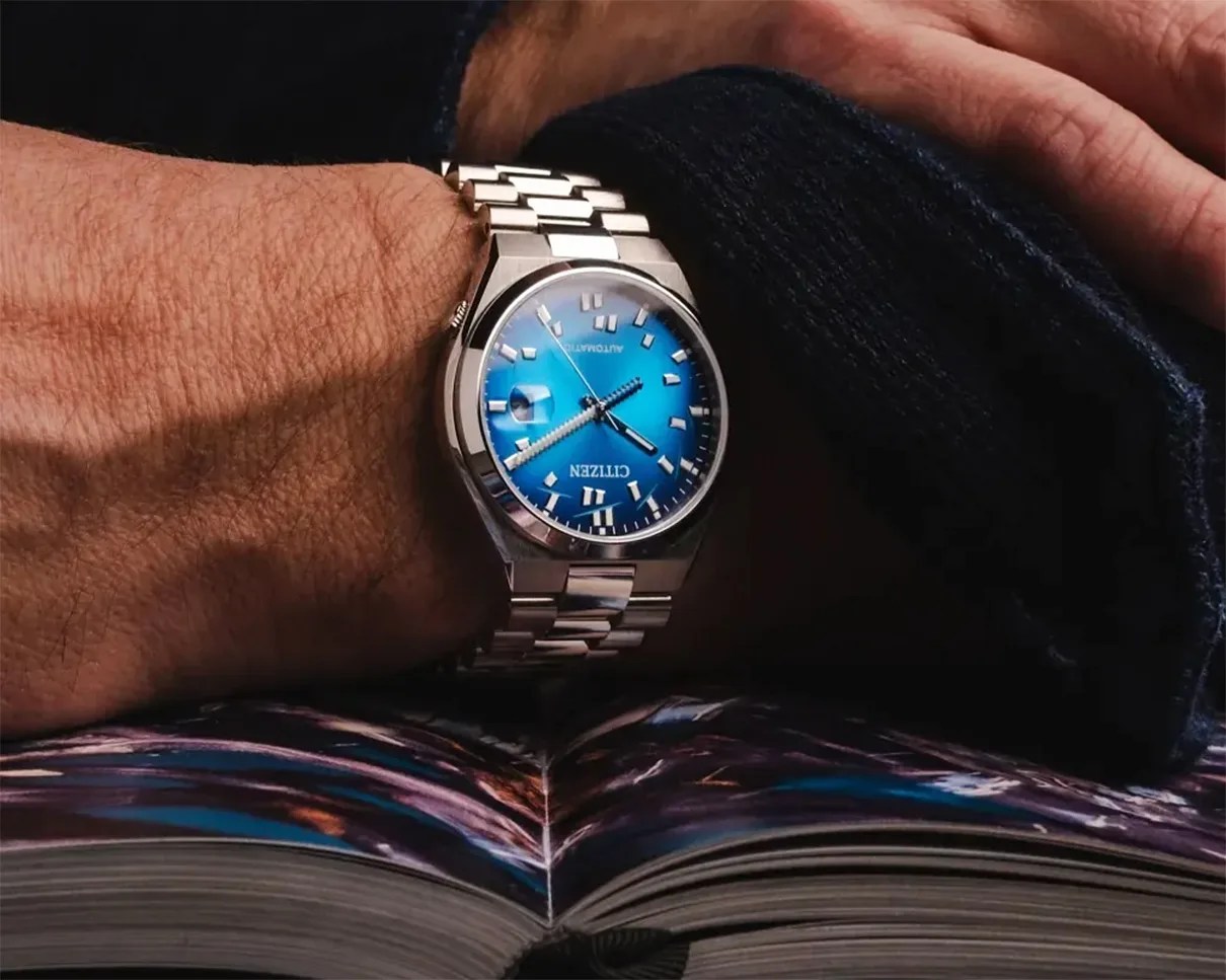 Silver Citizen wristwatch with a blue dial and metal link band worn on a wrist over an open book.