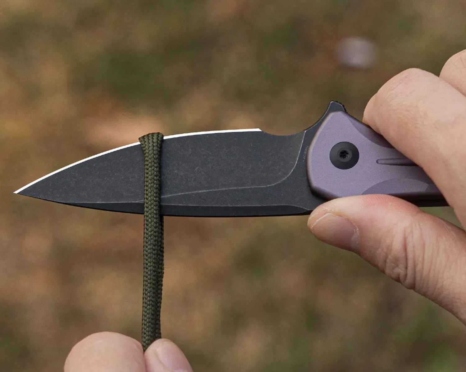 Hand holding a black stonewashed knife blade with a purple handle and green paracord looped around the blade.