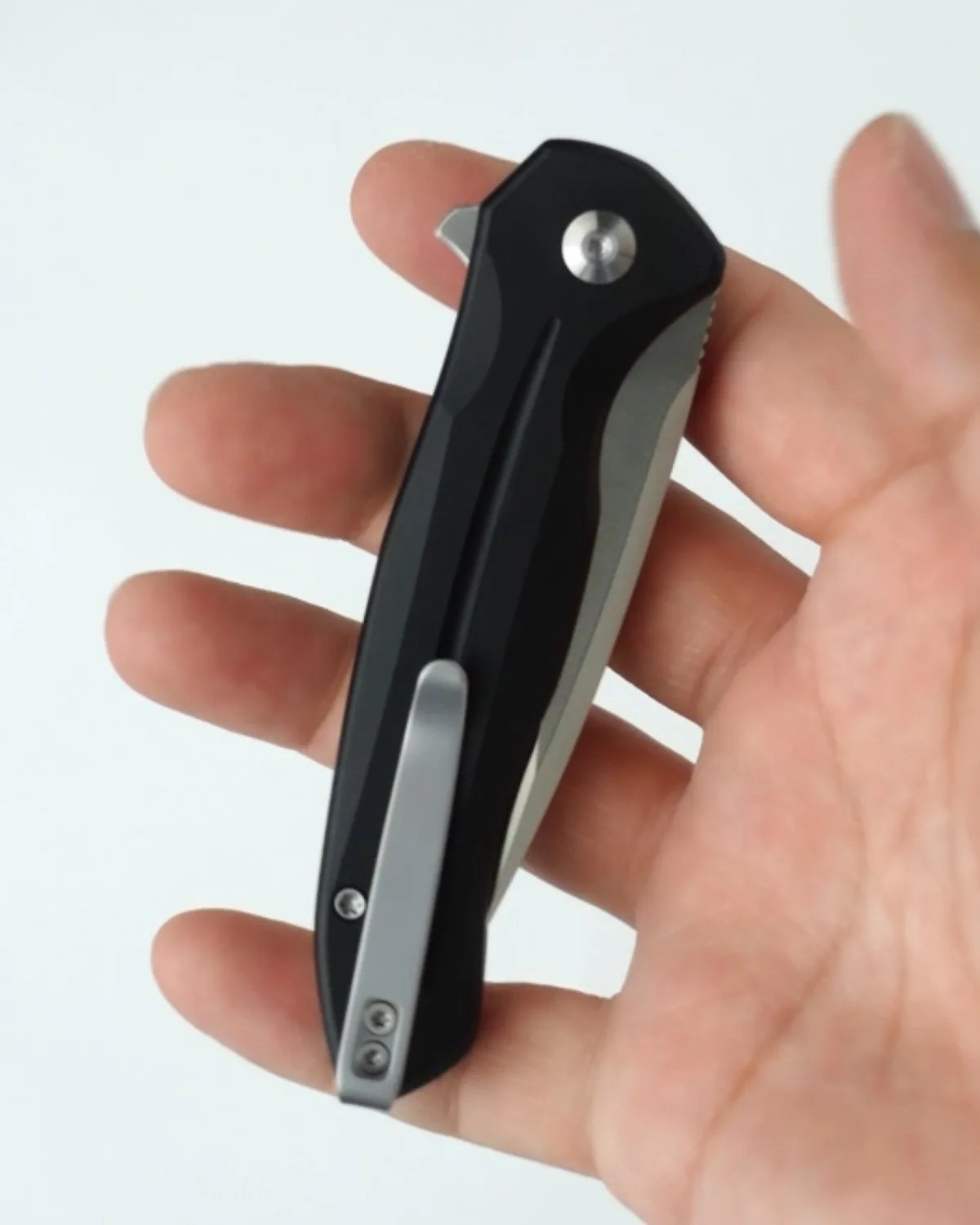 Black folding pocket knife with silver clip held in a hand against a white background.