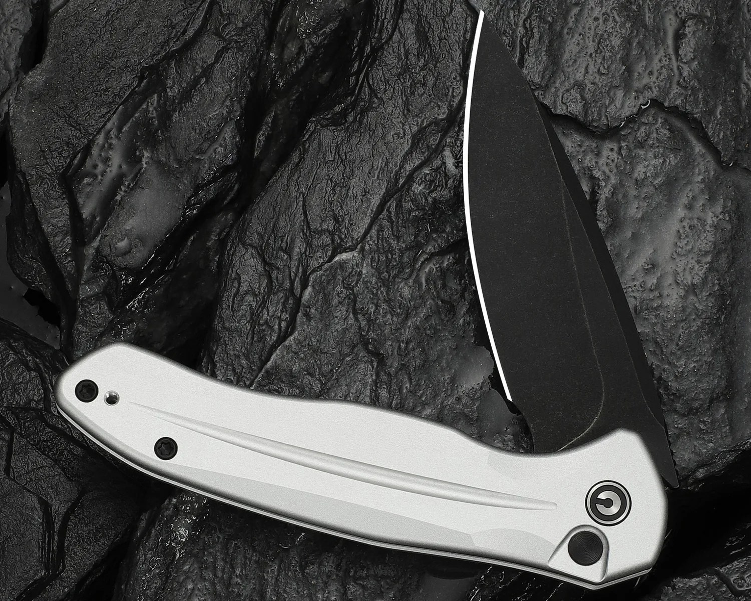 Folding knife with a matte black blade and a silver handle resting on textured black rocks.