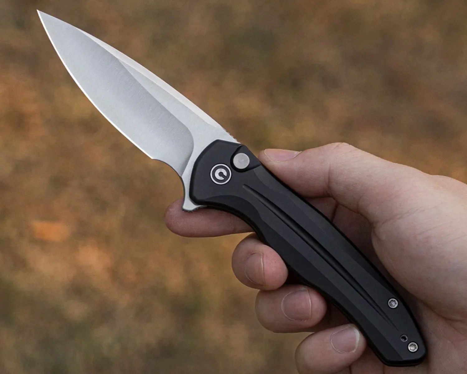 Hand holding a folding knife with a silver blade and black handle featuring a circular logo near the pivot.