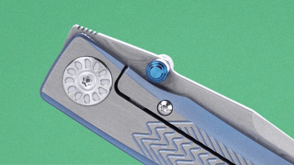 Close-up of a silver folding knife with a blue thumb stud and textured handle against a green background.