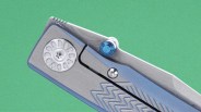 Close-up of a silver folding knife with a blue thumb stud and textured handle against a green background.