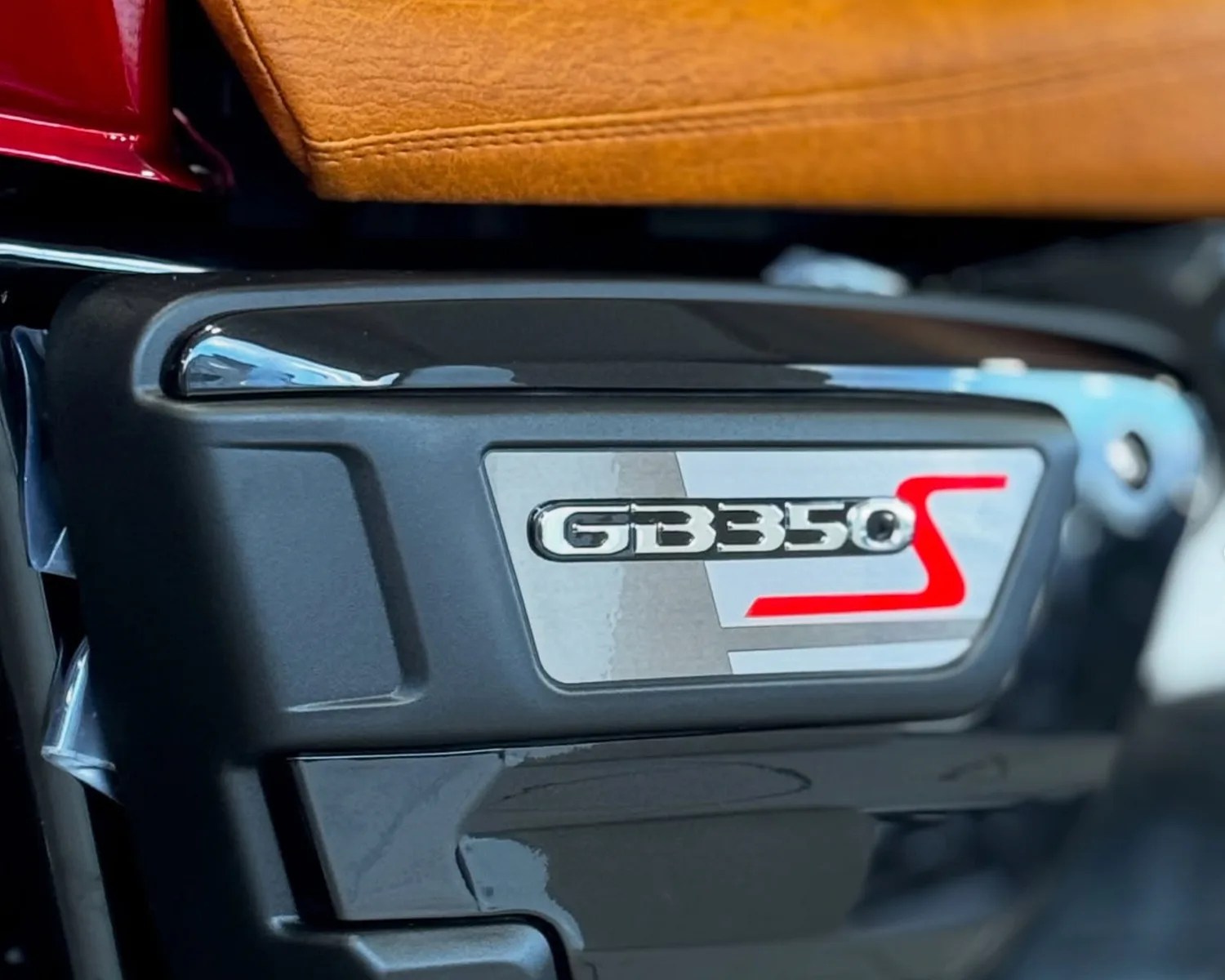 Close-up of a GB350S motorcycle emblem on a black plastic panel below a brown leather seat.